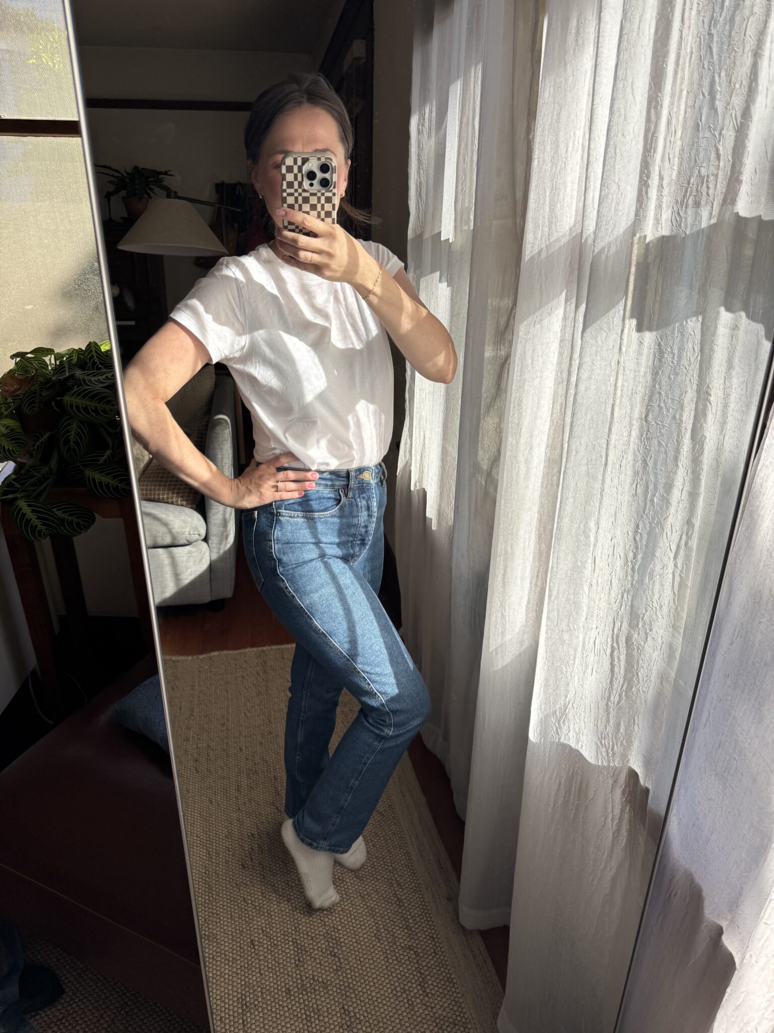 Person stands in front of a mirror taking a selfie, wearing a white t-shirt, blue jeans, and white socks, with sunlight streaming through sheer curtains.