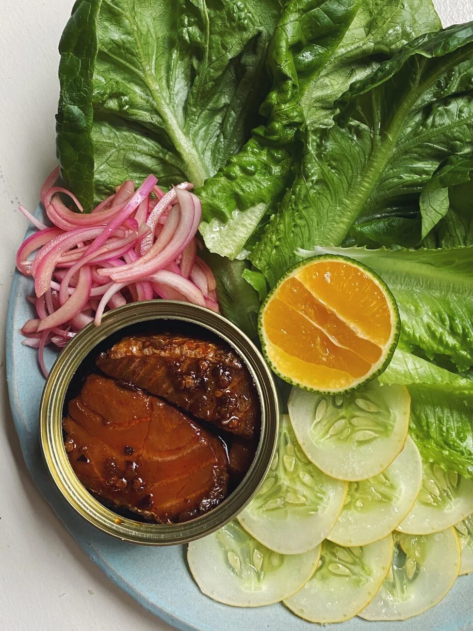 A plate with romaine lettuce leaves, sliced cucumber, pickled red onions, a halved lemon, and an open tin of fish in sauce.