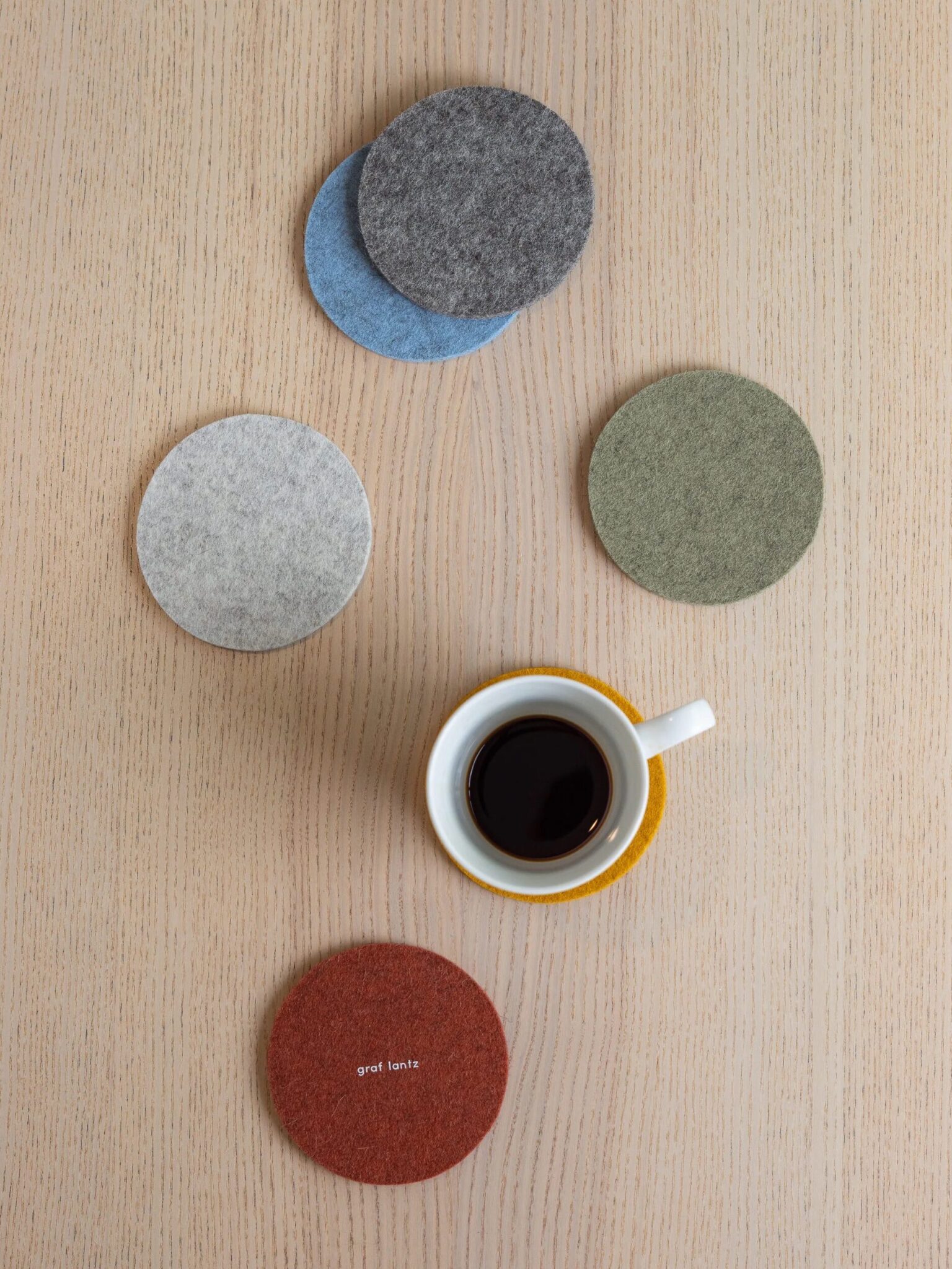 A cup of black coffee sits on a wooden table surrounded by five round felt coasters in gray, blue, green, and red colors.