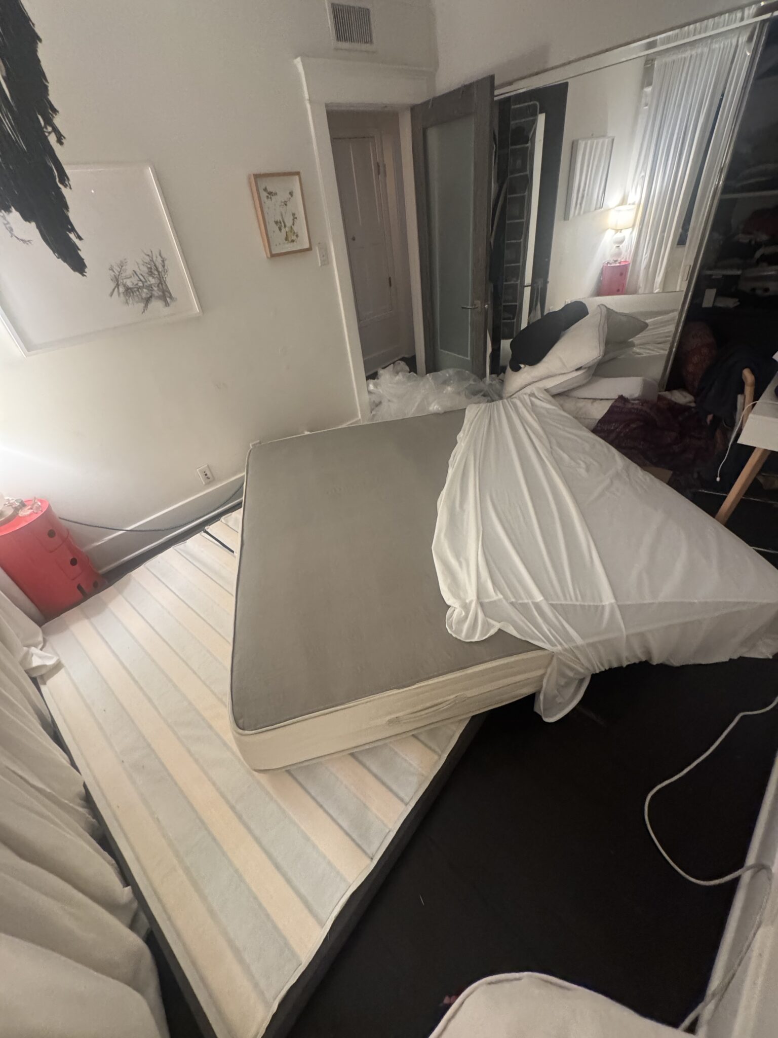 A mattress partially off its bed frame lies askew in a messy bedroom, with bedding and pillows displaced and furniture visible in the background.