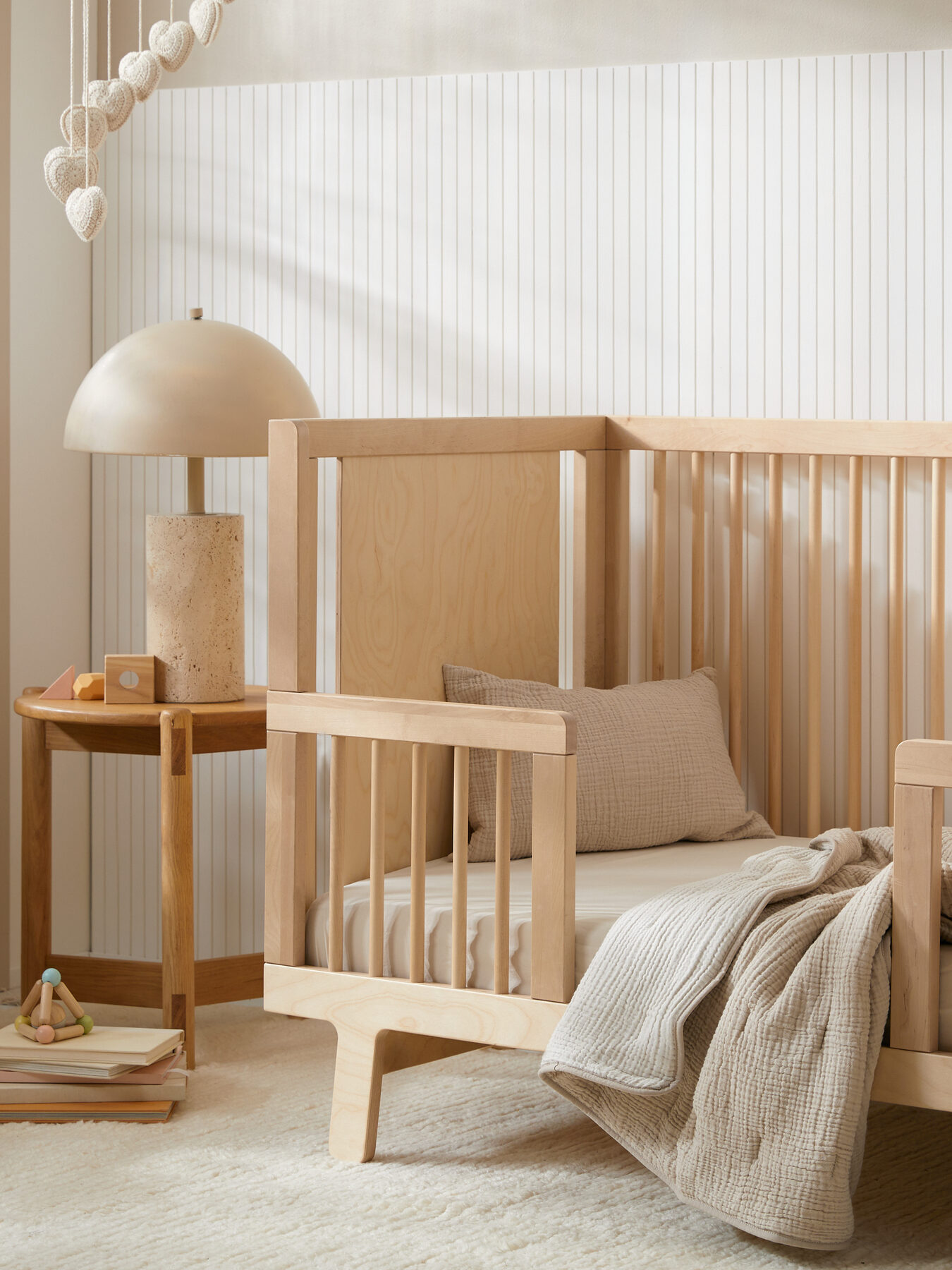 A modern nursery with a light wood crib, beige bedding, a wooden side table with a lamp, toys, and neutral decor on a textured rug.