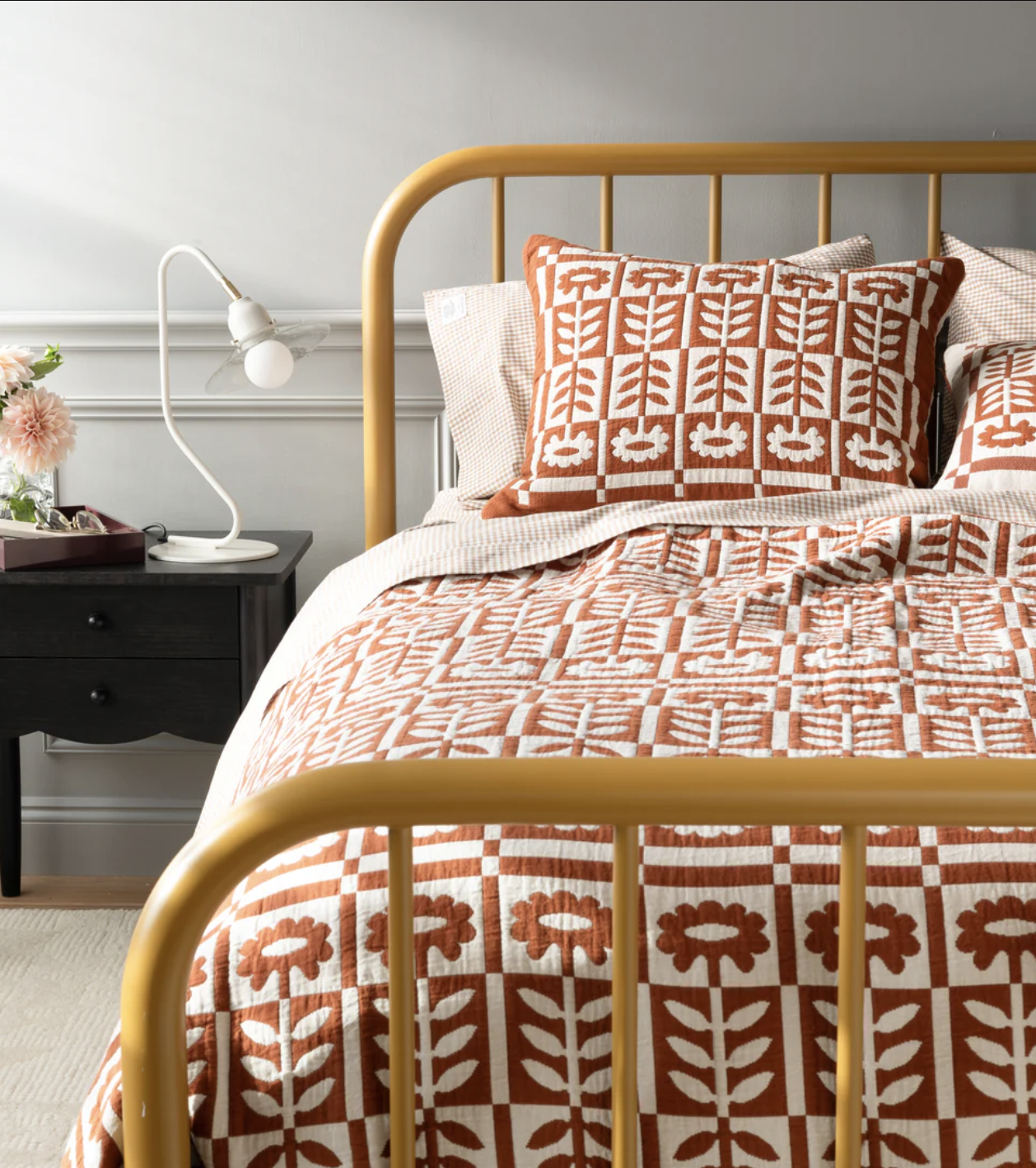 A neatly made bed with a mustard yellow metal frame, featuring a patterned orange and white quilt and pillow, next to a black nightstand with a white lamp and a pink flower.