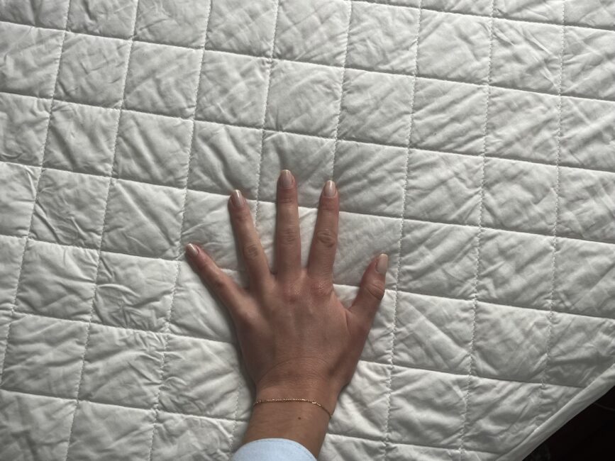 A left hand with light skin and a bracelet rests on a white, quilted fabric surface featuring a square pattern.