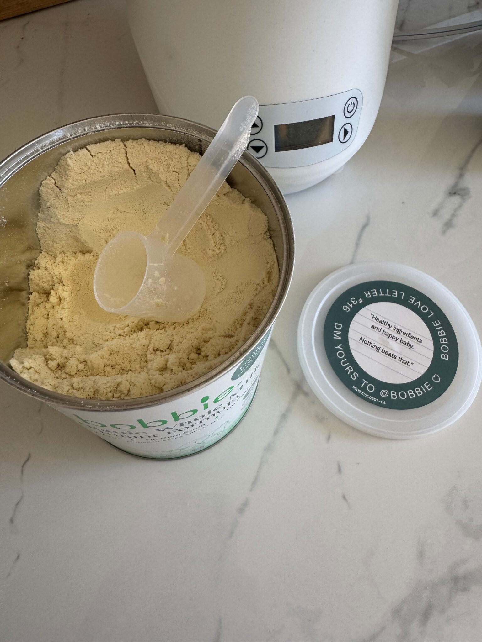An open container of powdered formula with a scoop inside sits on a marble countertop next to its lid and a white electronic appliance.