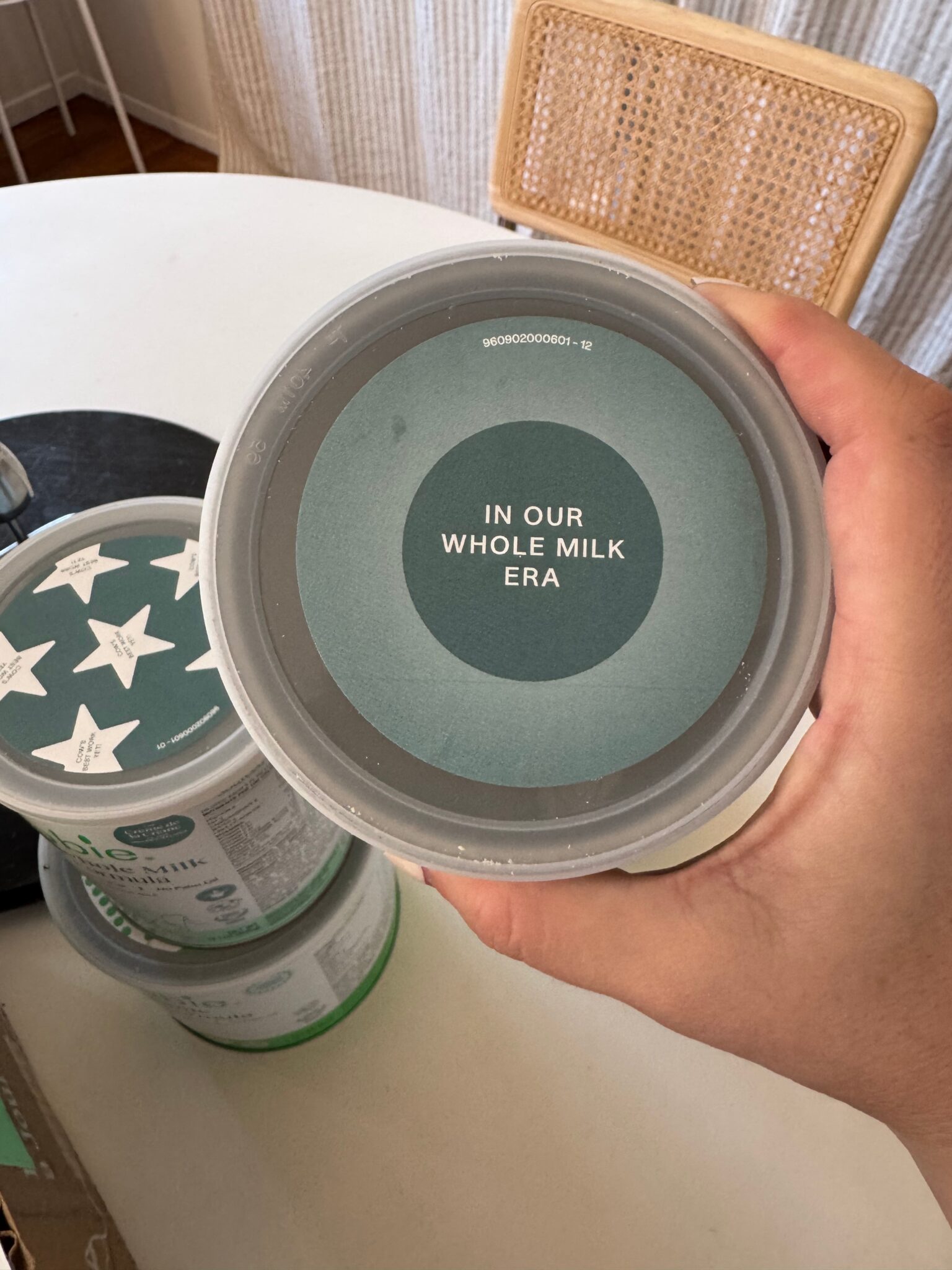 A hand holds a container with a lid labeled "IN OUR WHOLE MILK ERA." Other similar containers are visible on the table in the background.
