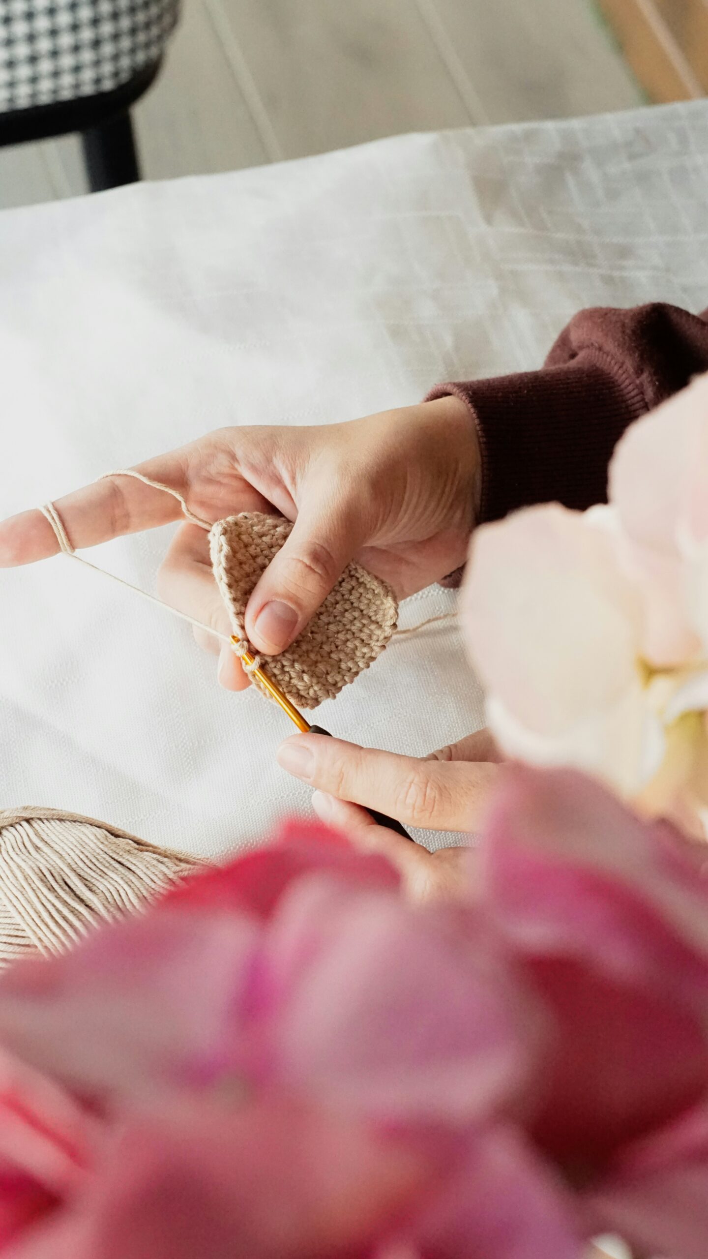 A person crocheting with beige yarn, holding a crochet hook and a small crochet piece, surrounded by blurred flowers in the foreground.