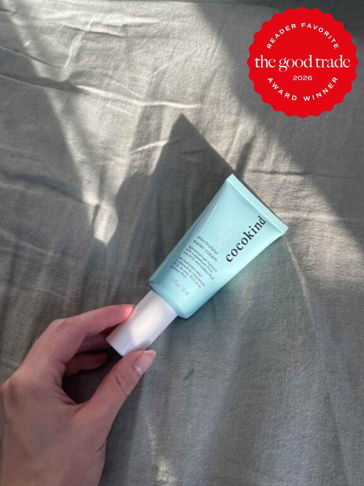 A hand holds a tube of Cocokind moisturizer on a gray fabric surface; a red badge in the corner reads “the good trade award winner, reader favorite 2026.”.