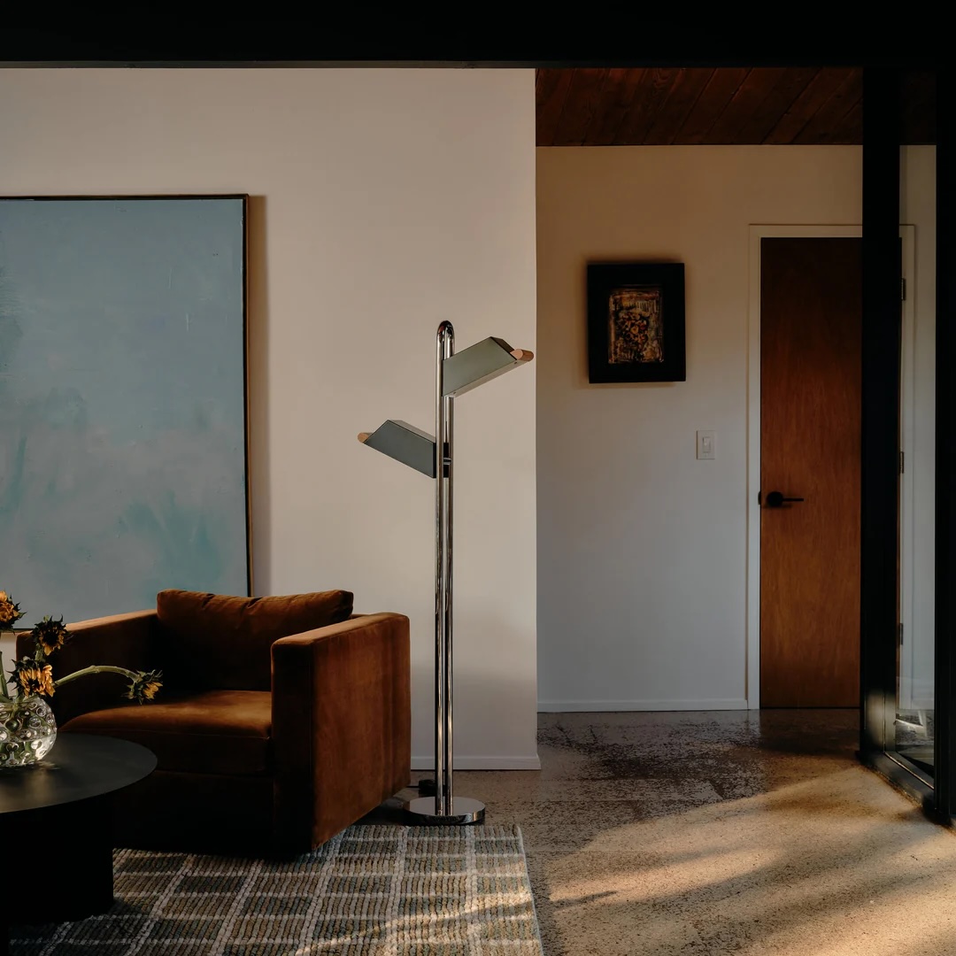 A modern living room with a brown armchair, a floor lamp with two adjustable shades, a large abstract painting, and a wooden door. Sunlight streams onto the patterned rug.