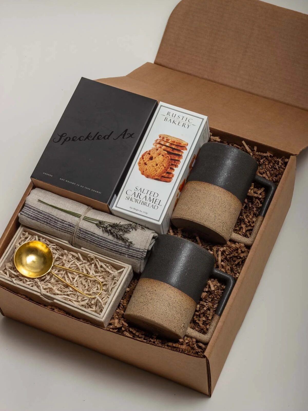 A gift box containing two ceramic mugs, a small gold spoon, a folded cloth, salted caramel shortbread cookies, and a box labeled "Speckled Ax", arranged on shredded paper.