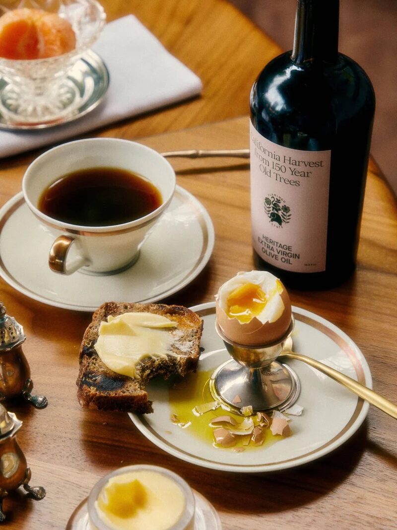 A breakfast table with a cup of black coffee, buttered toast, a soft-boiled egg with olive oil, and a bottle labeled “California Harvest Extra Virgin Olive Oil.”.