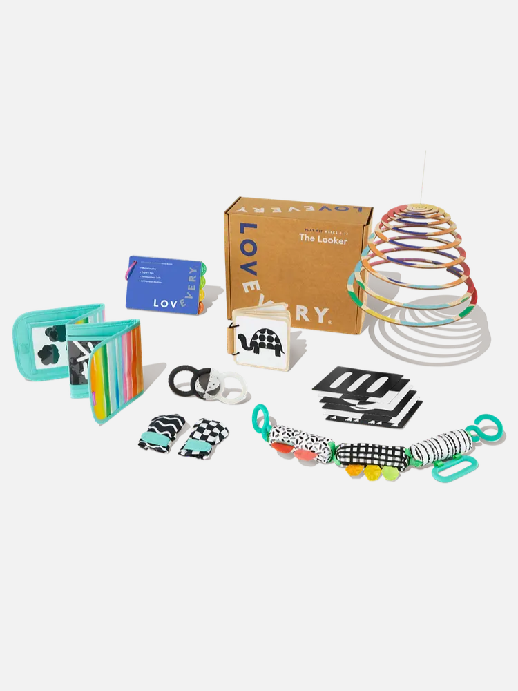 A set of colorful baby toys and flashcards displayed in front of a brown Lovevery box, including a rainbow slinky, soft books, black-and-white cards, and teething rings.