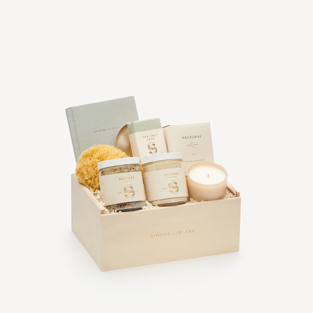 A wooden gift box containing a candle, jars, sponge, soap, book, and packaged items, all arranged neatly on crinkle paper.