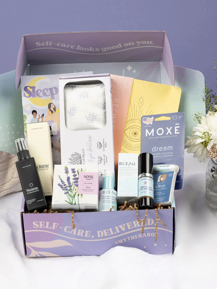 An open self-care subscription box displays various wellness products, including sprays, creams, supplements, eye pillows, and booklets, arranged neatly against a purple background.