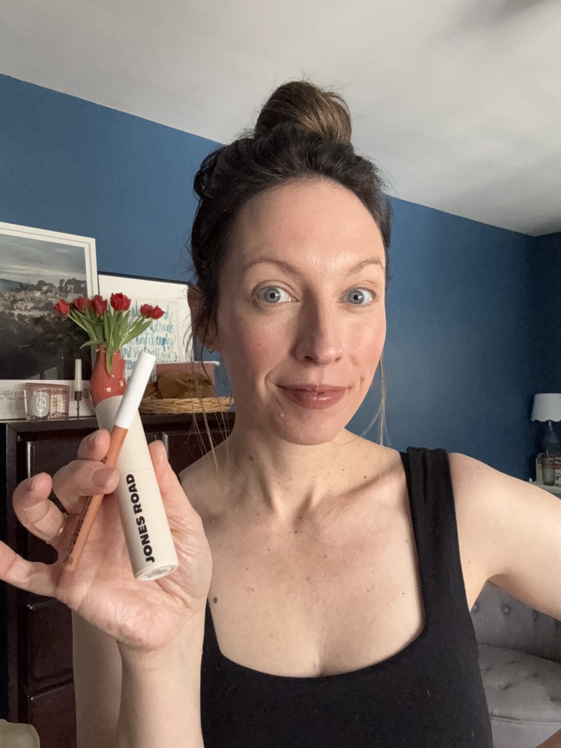 Woman with light skin and blue eyes, wearing a black tank top, smiles and holds up a Jones Road or Ilia beauty product in a room with blue walls and a vase of red tulips.