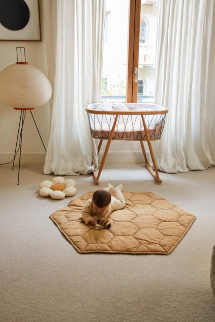 A baby lies on a tan hexagonal mat in a carpeted room with white curtains, a bassinet, a floor lamp, and a plush toy.