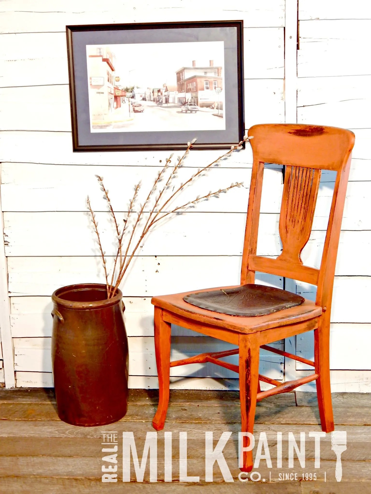 A rustic wooden chair sits next to a brown ceramic crock with dried branches, beneath a framed street scene, against white paneled walls. "The Real Milk Paint Co." logo is in the corner.