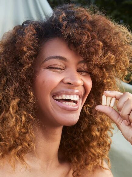 A person with curly hair smiles and holds three supplement capsules between their fingers in an outdoor setting.