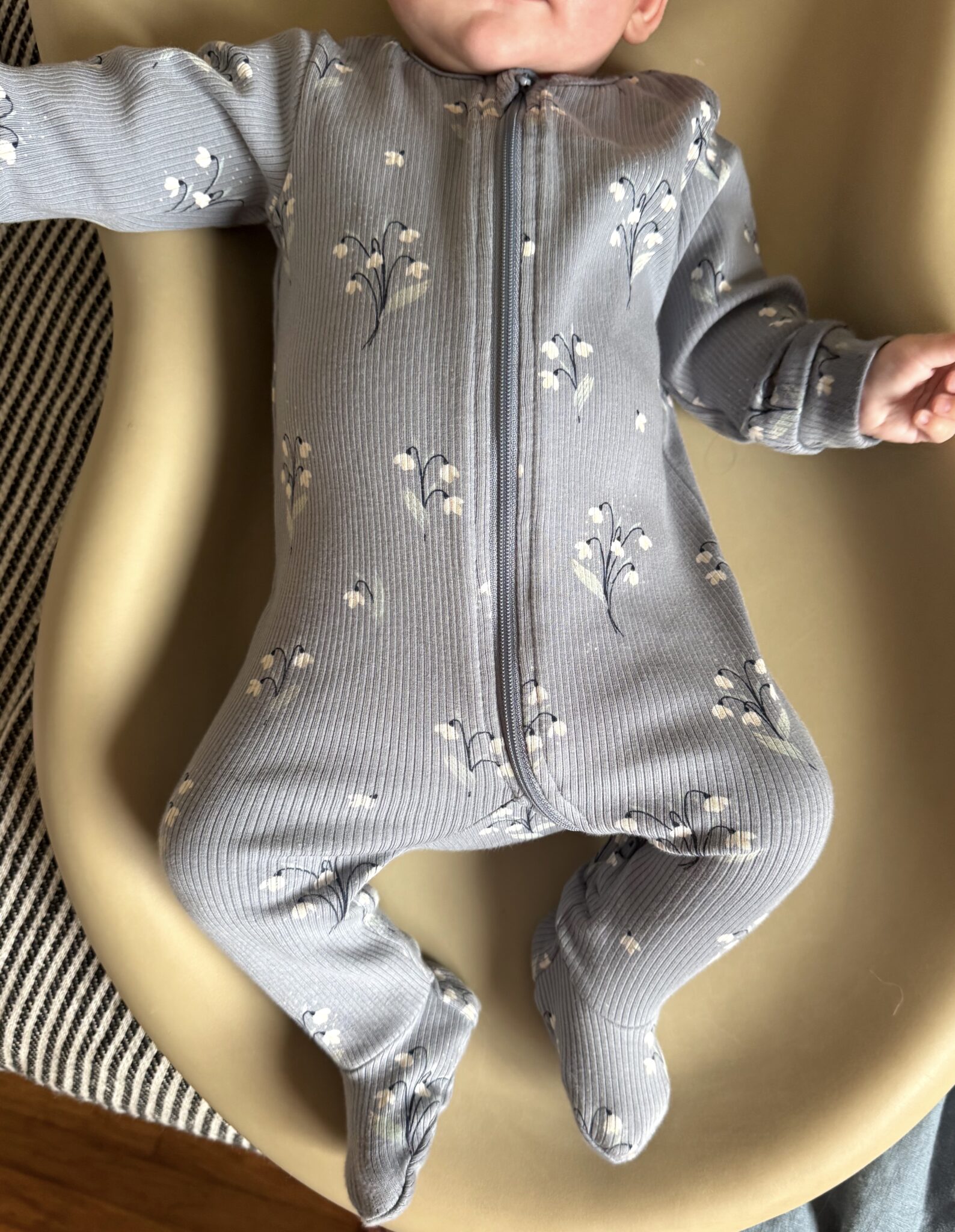 A baby in a gray, floral-patterned zip-up onesie lies on a beige changing pad, with part of their face out of frame.