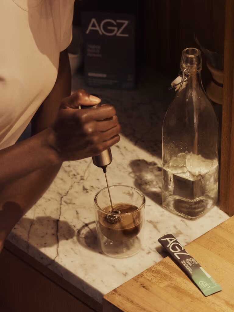 A person uses a handheld frother to mix a drink in a glass on a marble countertop, with an AGZ packet and a glass bottle nearby.