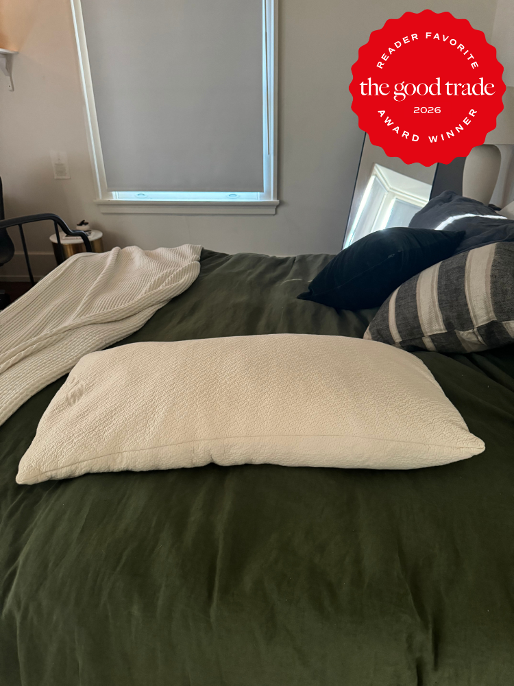 A neatly made bed with a green duvet, a white pillow in the center, and a white blanket partially draped. A “The Good Trade Award Winner 2026” badge appears in the upper right corner.