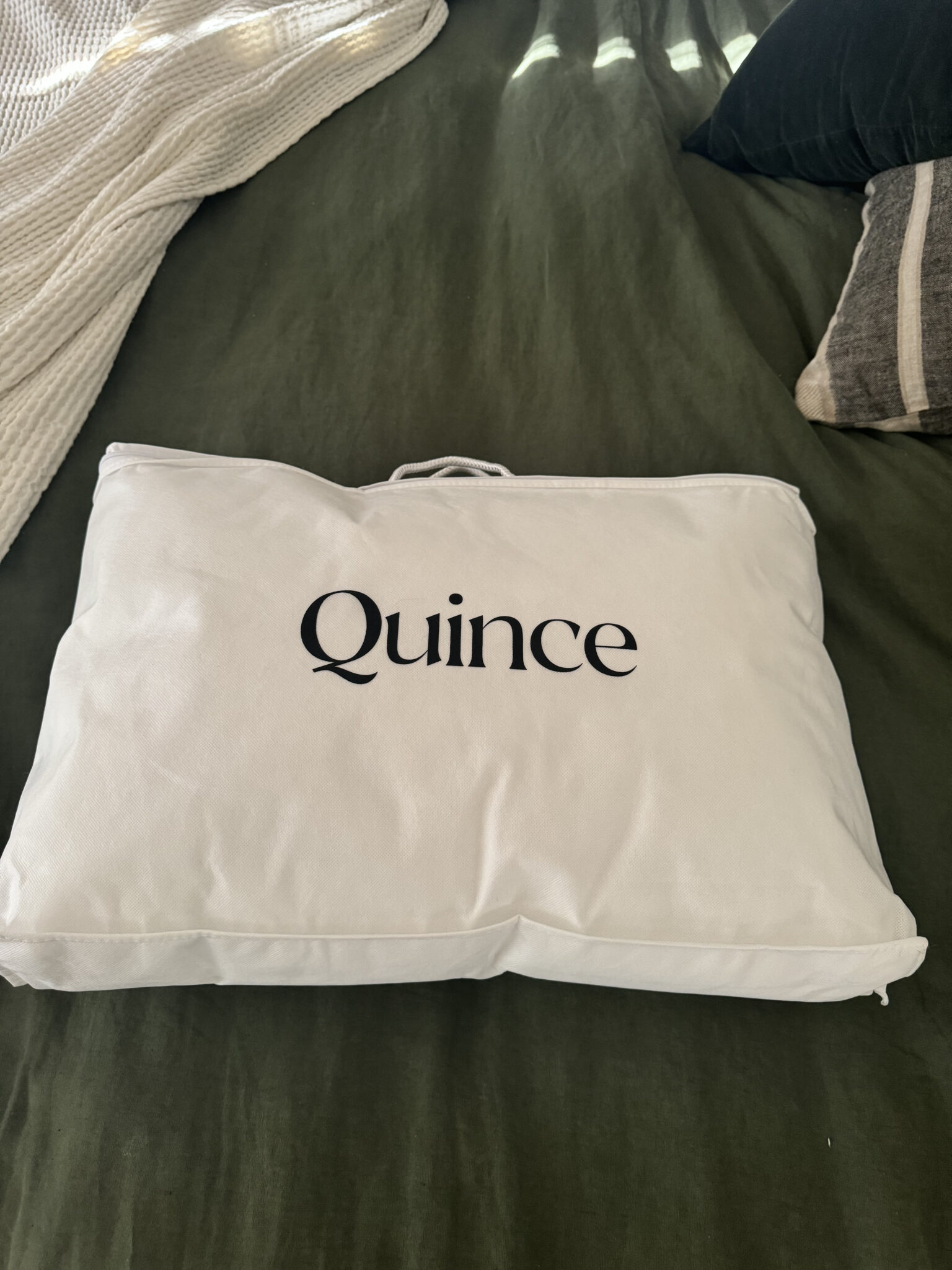 A white Quince-branded fabric bag rests on a green bedspread with pillows and a cream knit blanket nearby.