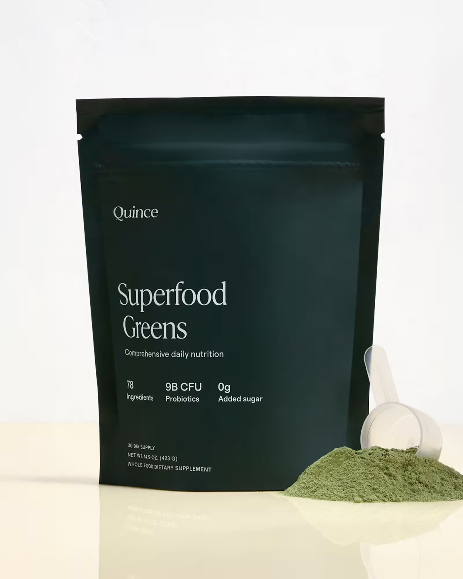 A black pouch labeled "Quince Superfood Greens" stands upright with a clear scoop and a small pile of green powder next to it on a light surface.
