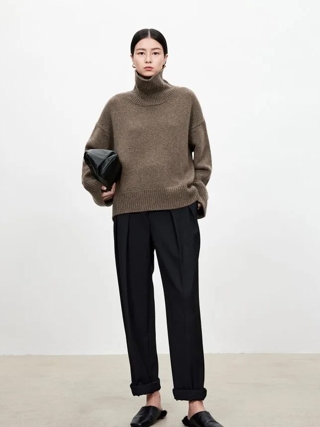 A person stands against a plain background wearing a brown turtleneck sweater, black pleated trousers, black slip-on shoes, and holding a black clutch.