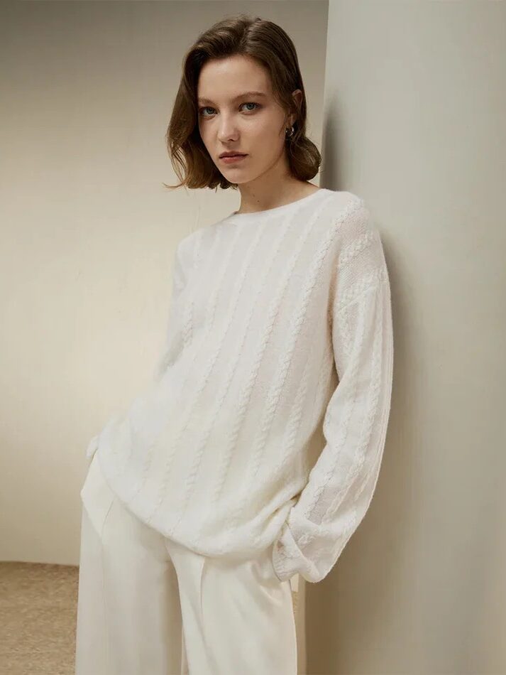 A person wearing a white textured sweater and white pants stands indoors, leaning against a curved wall, looking directly at the camera.