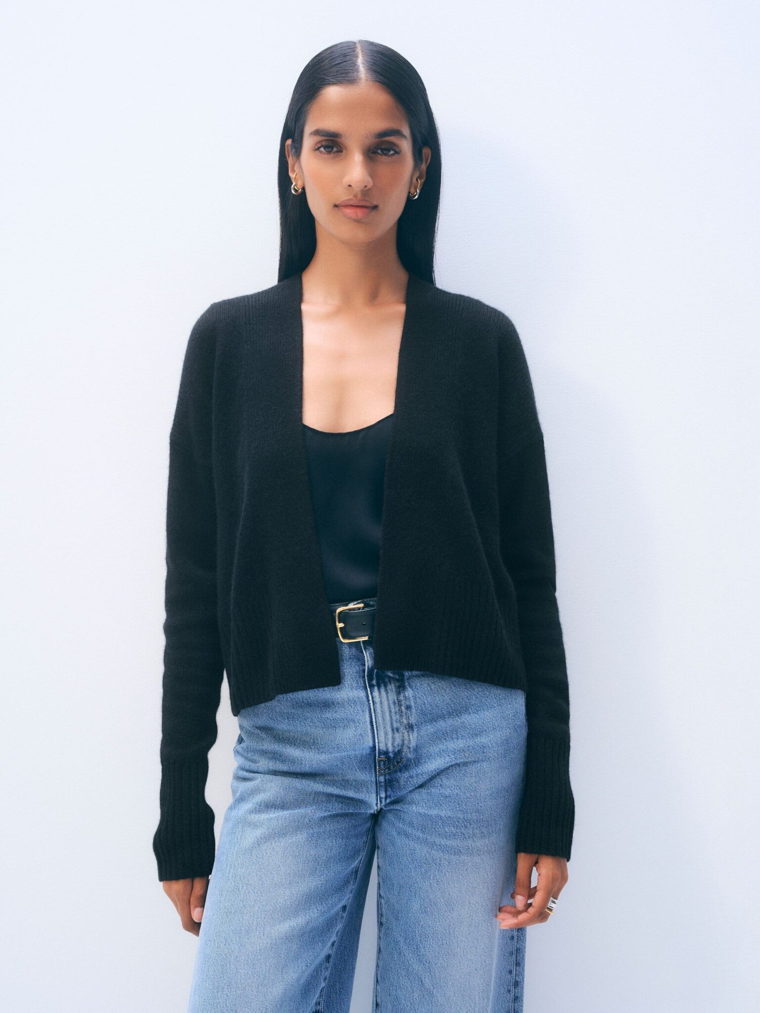 A woman stands against a plain light background, wearing a black cardigan over a black top, blue jeans, and a black belt.