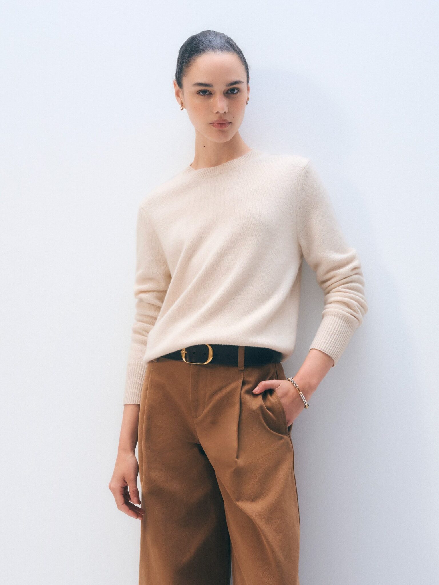 Person standing against a plain white background, wearing a beige sweater and brown trousers with a black belt, looking directly at the camera.