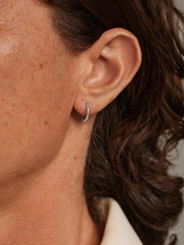 Close-up of a person's left ear wearing a small silver hoop earring, with brown hair visible and part of a cream-colored collar.