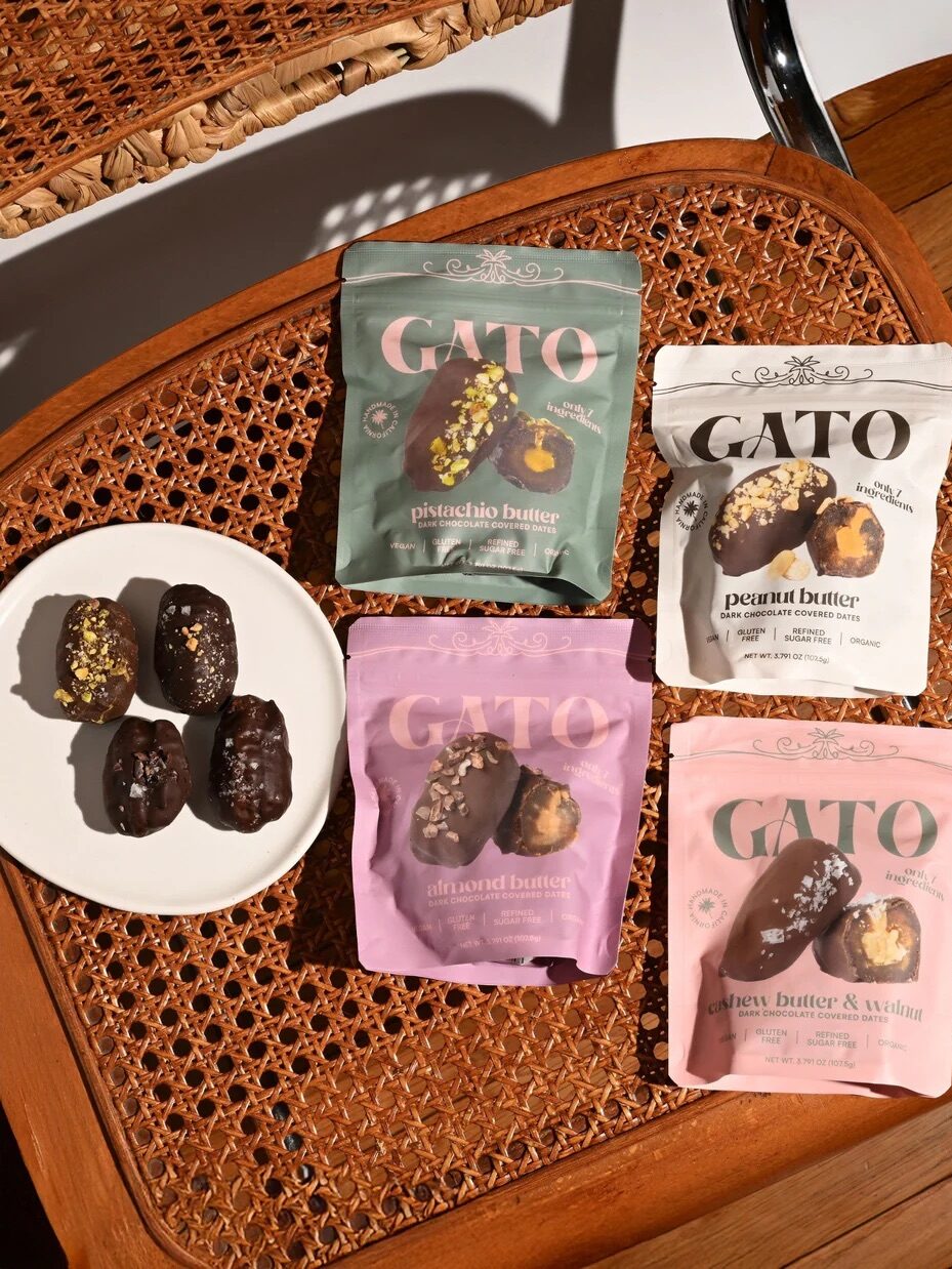 Four GATO snack pouches in different flavors are arranged on a woven chair next to a white plate holding five chocolate-coated snacks.