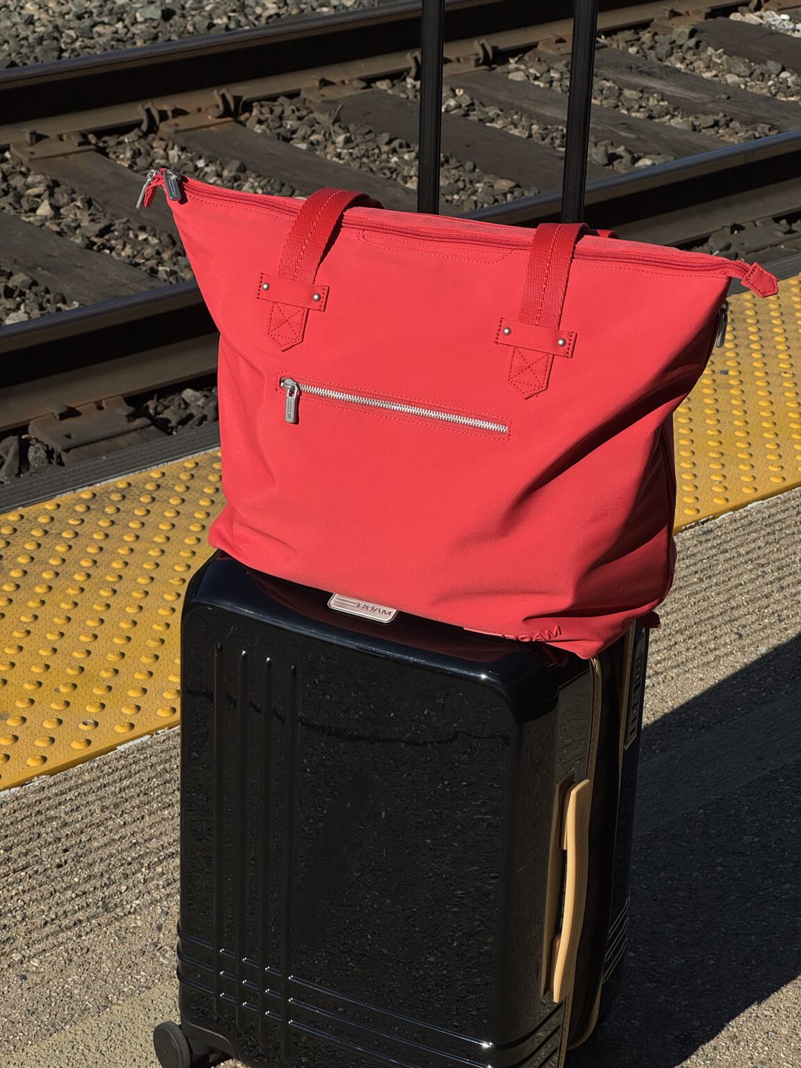 A red tote bag sits on top of a black rolling suitcase on a train platform near train tracks and a yellow tactile paving strip.