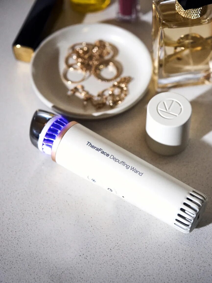 A TheraFace Depuffing Wand is placed on a bathroom countertop next to jewelry, a bottle of perfume, and a white cosmetic container.