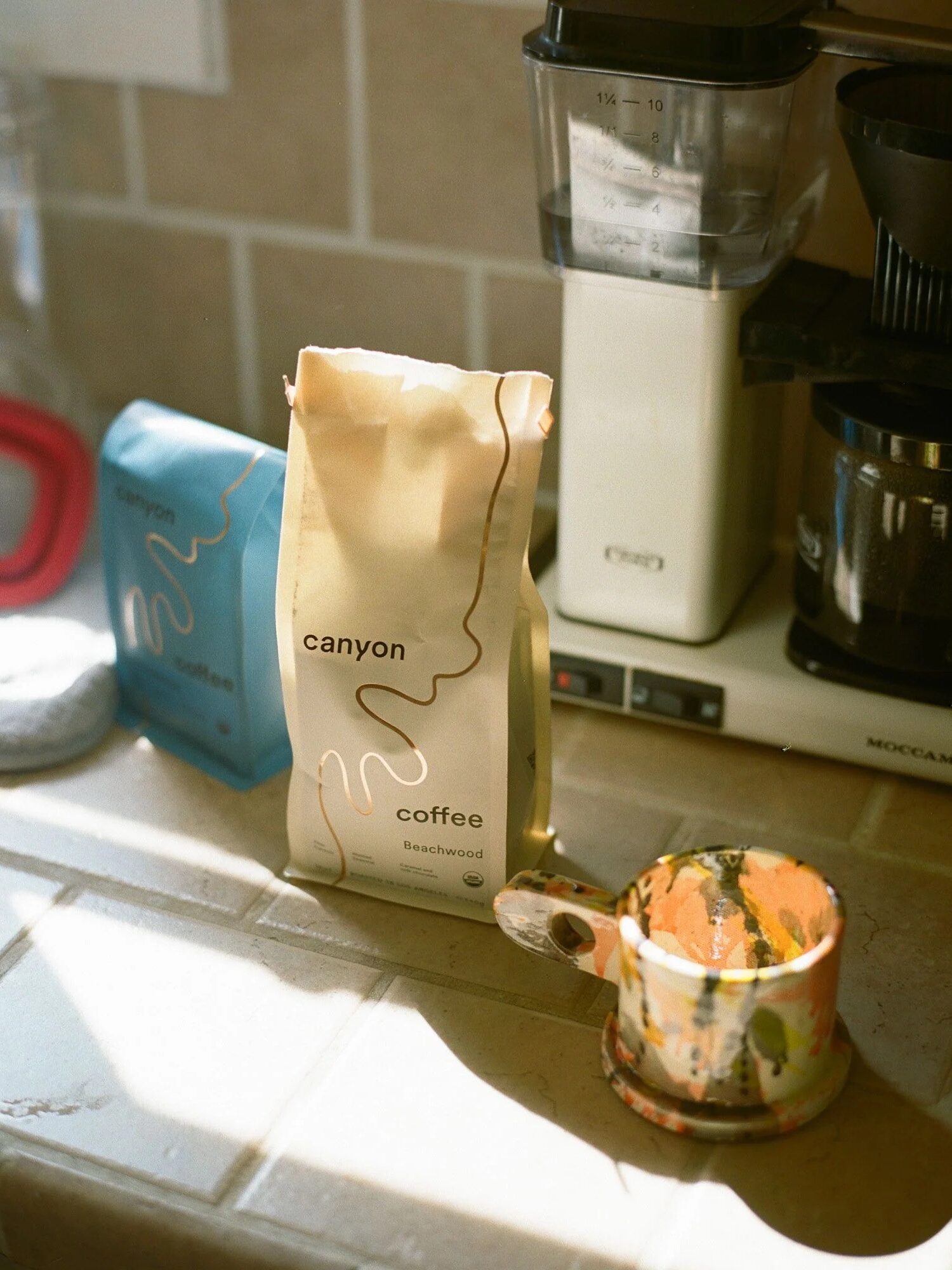 Bags of Canyon Coffee and a ceramic mug sit on a kitchen counter next to a coffee grinder and coffee maker in natural light.