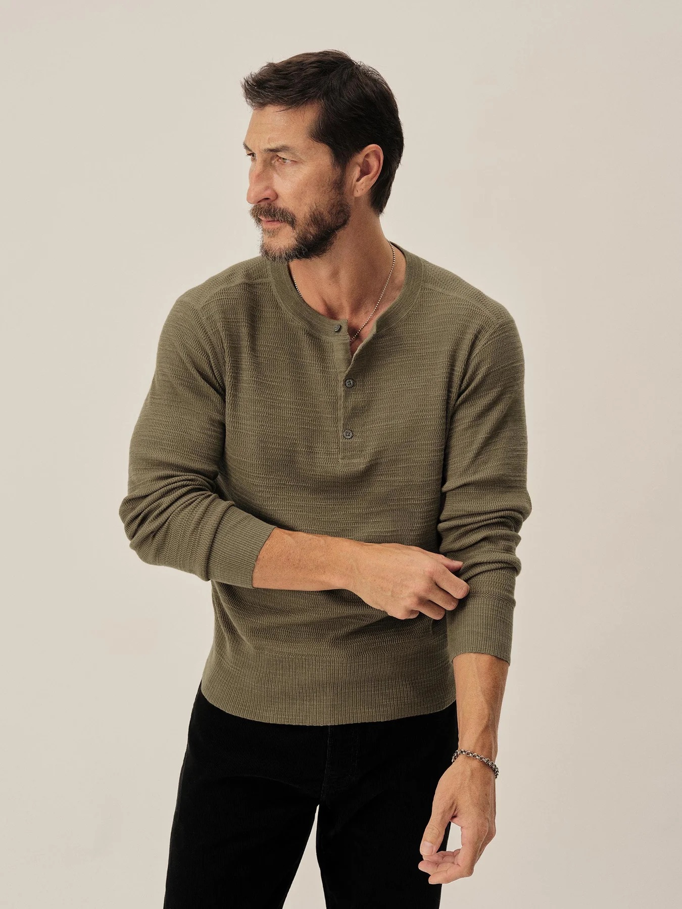 A man with a beard wearing a long-sleeve olive green henley shirt and black pants stands against a plain light background, looking to his left.