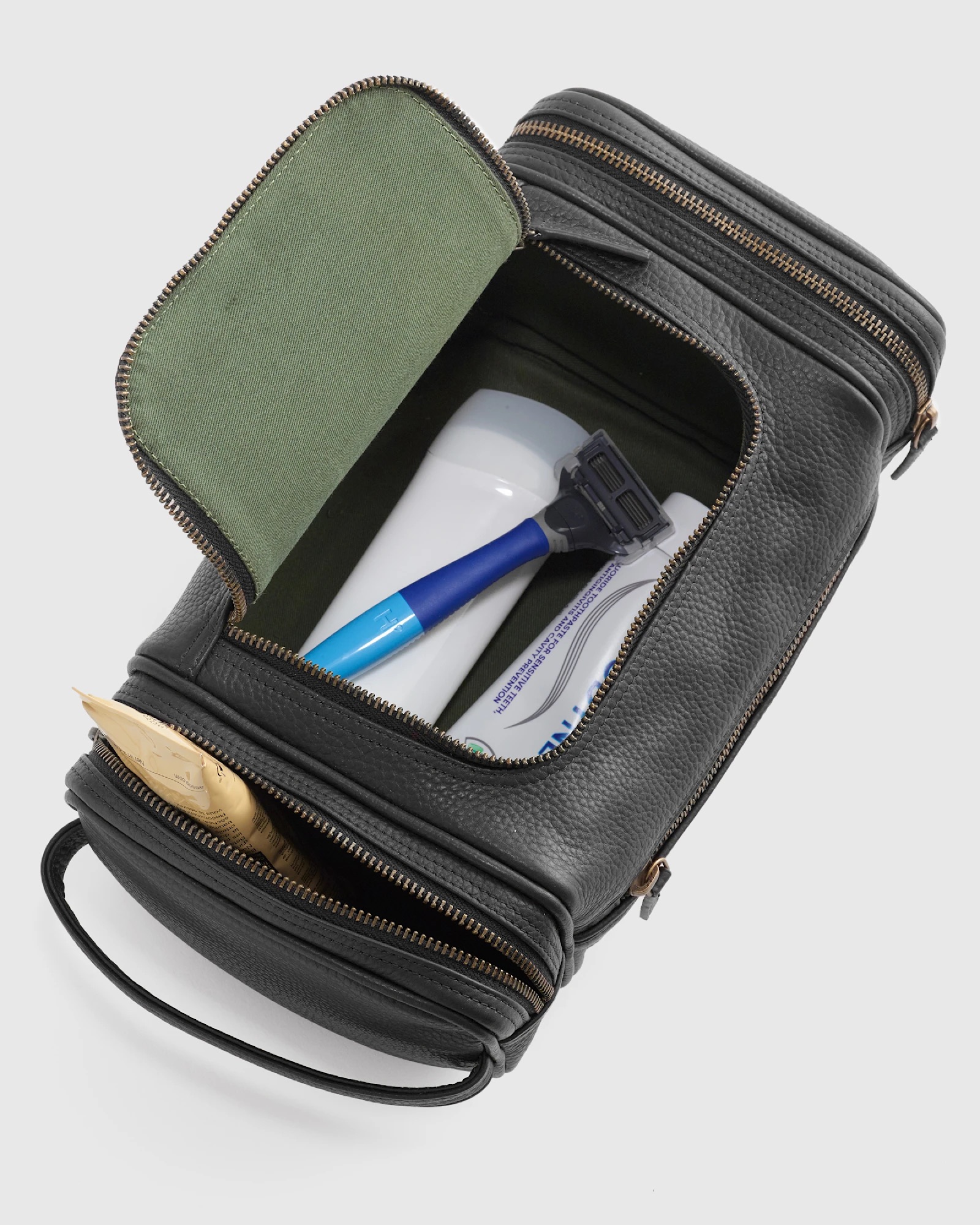 Black leather dopp kit with an open zipper showing a razor, toothpaste, and toothbrush inside; a packet is in the side pocket.