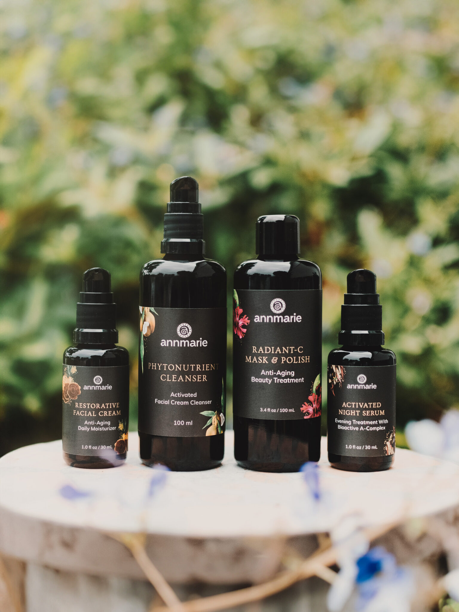 Four Annmarie skincare products—face cream, cleanser, mask, and night serum—are arranged on a wooden surface outdoors with greenery in the background.