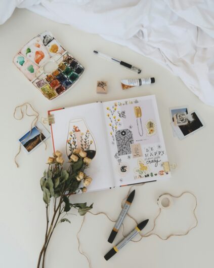 An open sketchbook with drawings and stickers lies on a desk surrounded by markers, dried flowers, watercolor paints, photos, string, and art supplies.