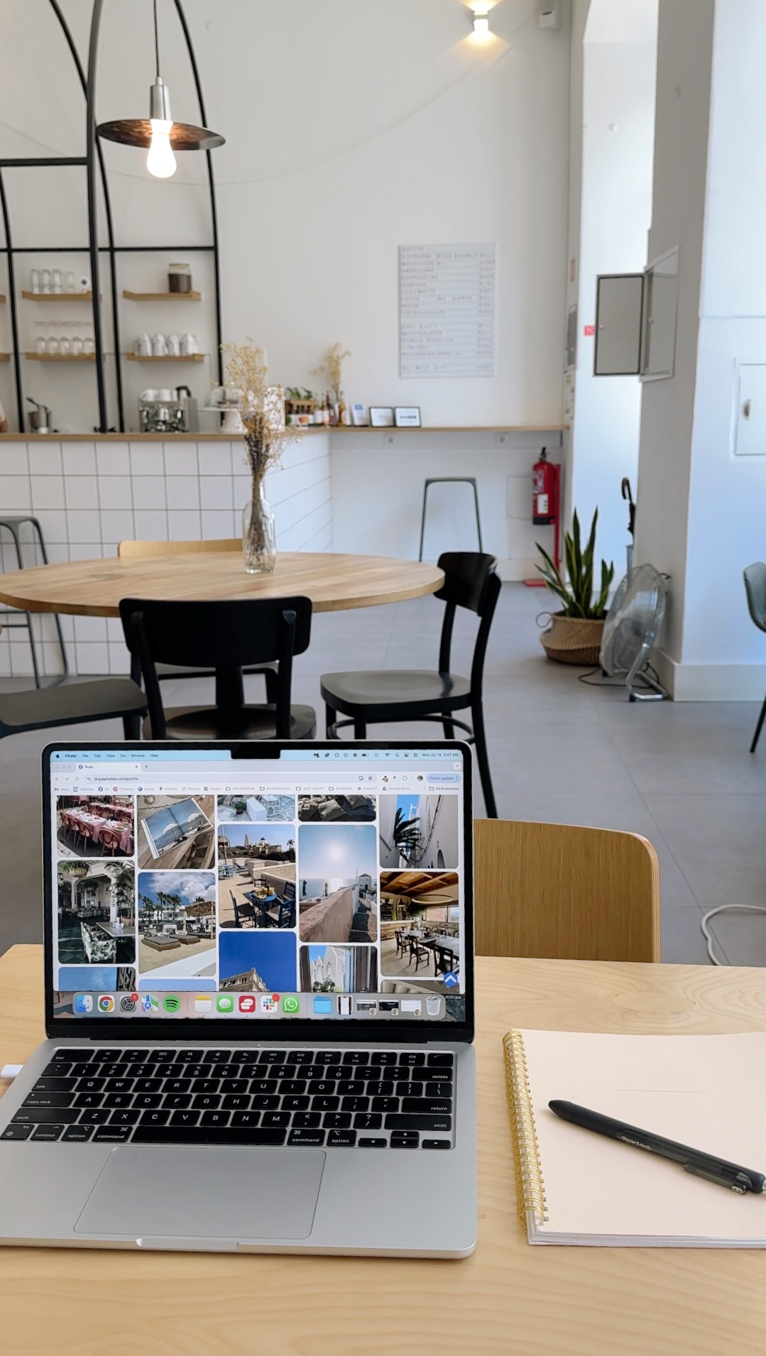 Open laptop on a table displaying interior photos; nearby, a notepad and pen, with a minimalist café setting and empty chairs in the background.