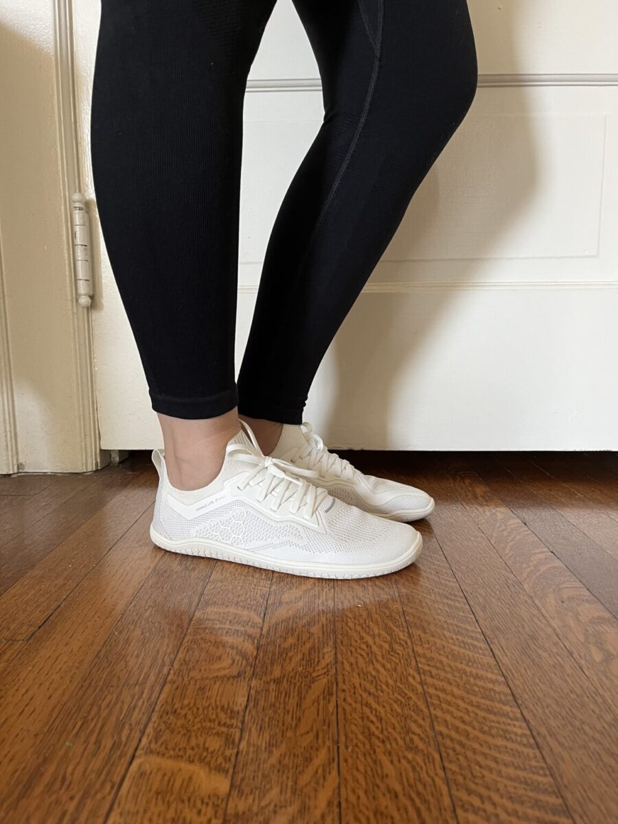 Person wearing black leggings and white sneakers standing on a wooden floor near a white door.