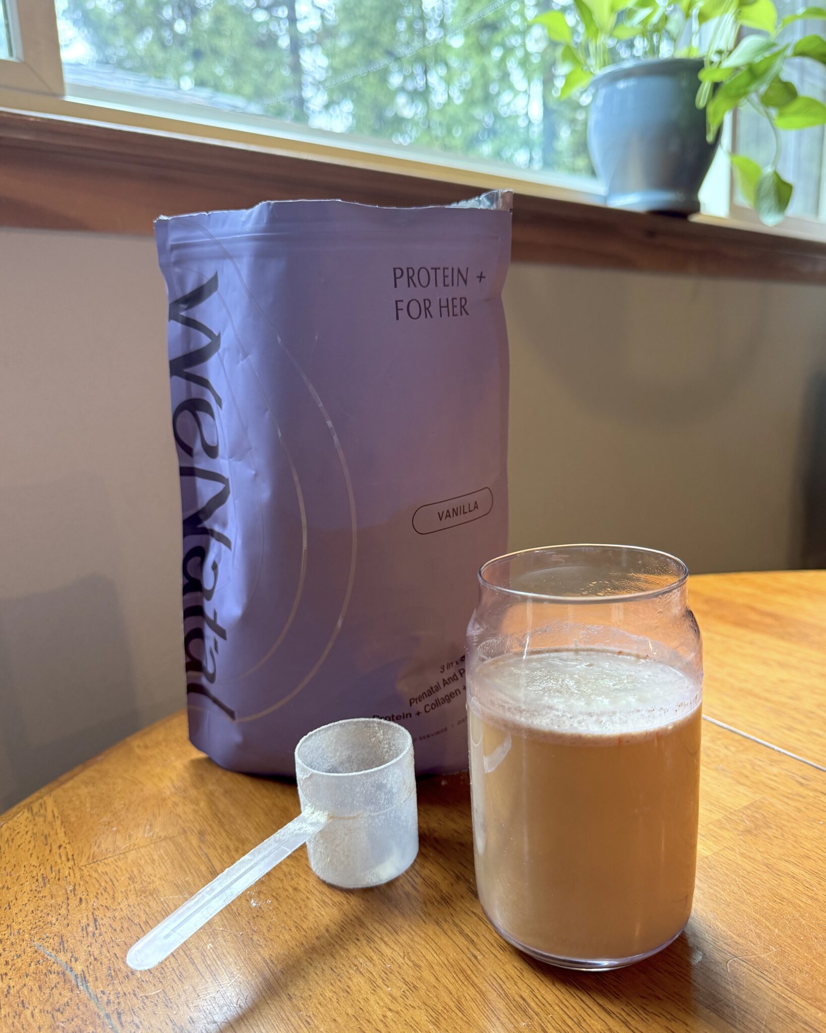 A bag of vanilla protein powder labeled "Protein + For Her" sits on a table with a clear scoop and a glass of mixed protein shake beside it. A window and plant are in the background.