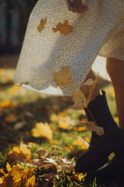 A person wearing a white patterned dress and black boots stands on grass covered with yellow autumn leaves.