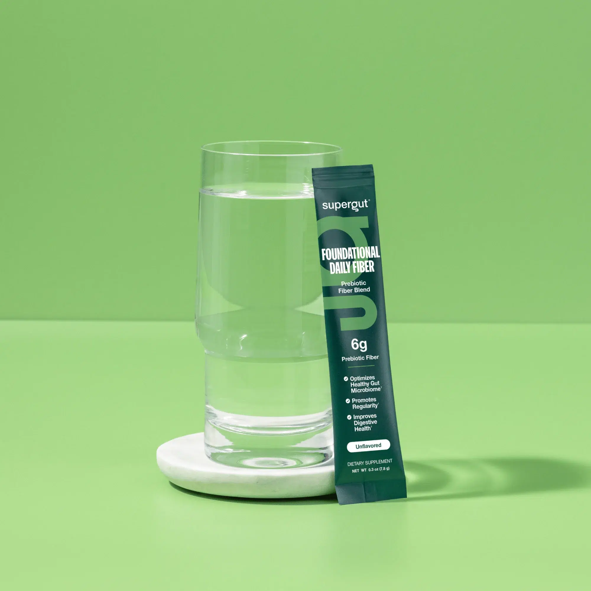 A glass of water on a white coaster sits beside a green packet labeled "Foundational Daily Fiber" by Supergut—an excellent choice among fiber supplements for women—set against a solid green background.