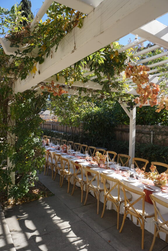 Long table set for an outdoor meal under a pergola with greenery and hanging floral arrangements; wooden chairs line both sides.