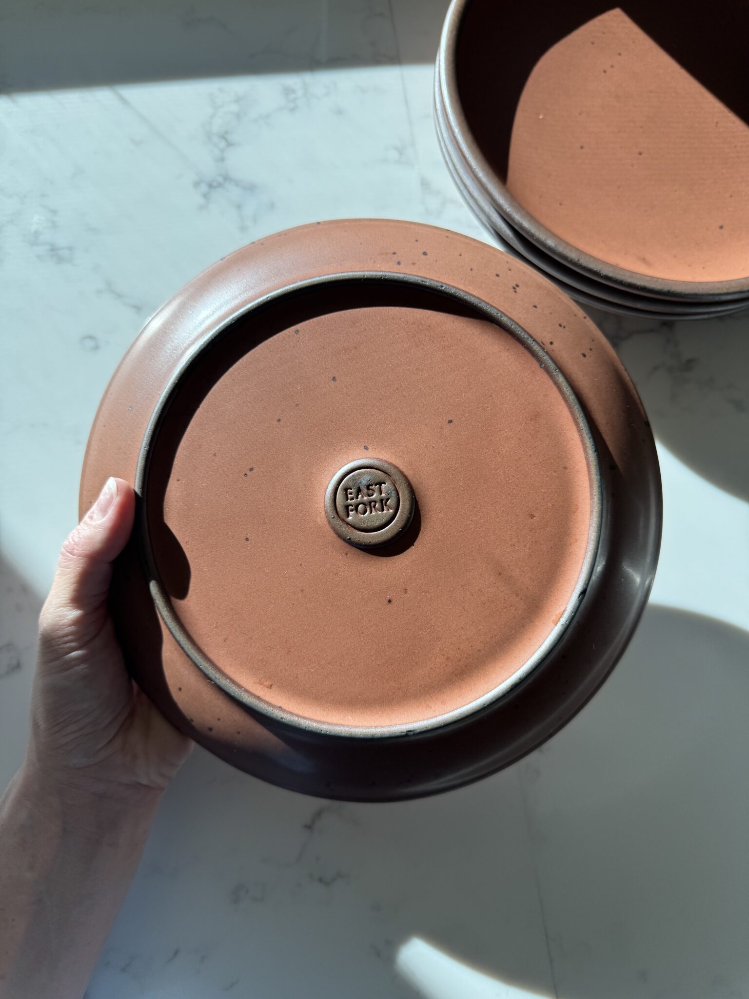 A hand holds an upturned ceramic bowl stamped "EAST FORK" on the base; another similar bowl is partially visible on a white surface.