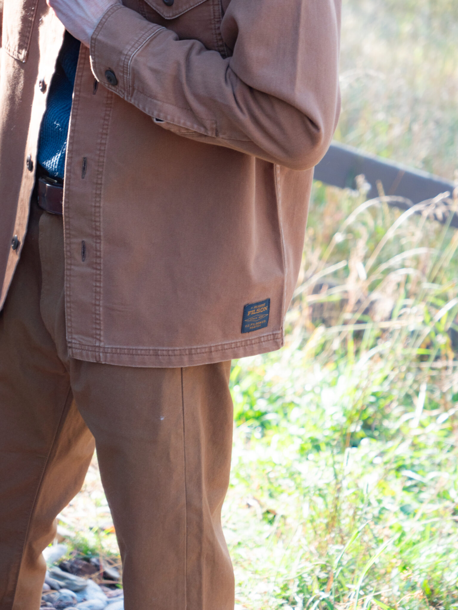 A person wearing a brown jacket and brown pants stands outdoors near green grass.