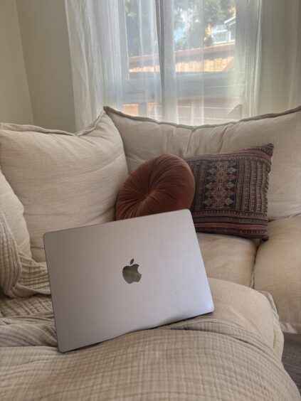 A closed silver MacBook sits on a beige couch with two decorative pillows, near a window with sheer curtains letting in natural light.