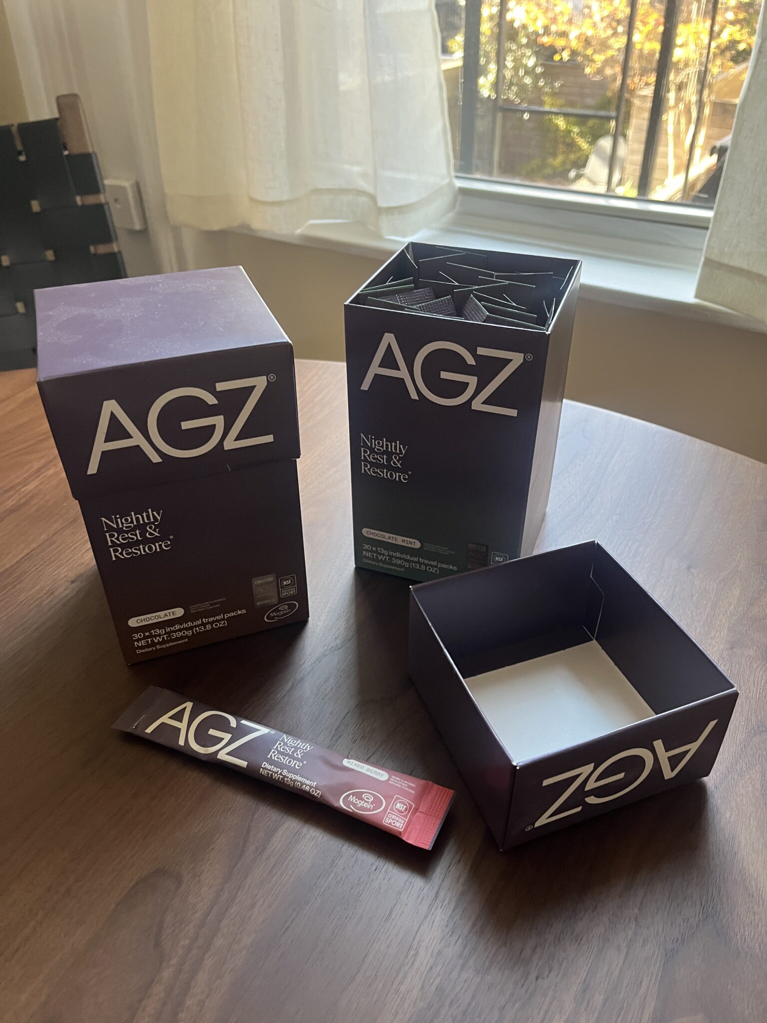 Two open boxes labeled "AGZ Nightly Rest & Restore" sit on a wooden table, with one single-serving stick pack placed in front of them. A window is in the background.