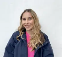 A person with long blonde hair, wearing a dark jacket over a bright pink top, stands in front of a plain white wall and smiles at the camera.